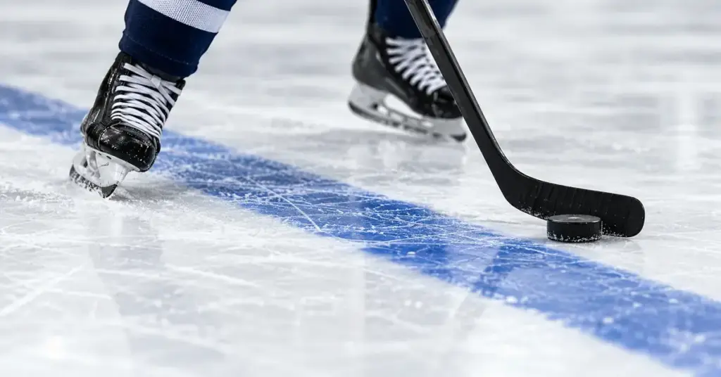 Jääkiekkoilijan luistimet, maila ja kiekko kaukalon siniviivan tuntumassa.